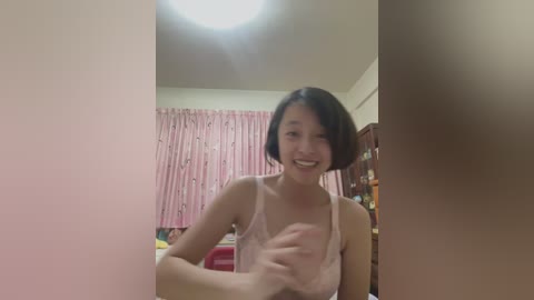 Video of a smiling East Asian woman with short black hair in a light pink camisole, standing in a pink-draped room with a white ceiling light.