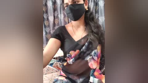 A video of a South Asian woman in a black floral saree with a face mask, sitting against a patterned curtain. She has long, dark hair and gold earrings.