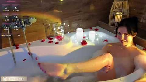 A video of a topless woman with fair skin and dark hair, reclining in a bathtub filled with bubbles and rose petals, surrounded by lit candles and a white curtain in a dimly lit, cozy room.