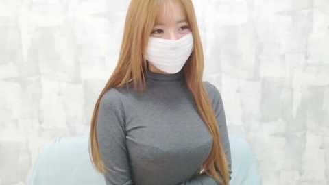 Video of a young Asian woman with long, straight, light brown hair, wearing a gray long-sleeve shirt, white surgical mask, and light blue chair, against a white, patterned background.