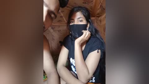Video of a South Asian woman with medium skin tone, long black hair, and a black face mask, wearing a black T-shirt with a white graphic, sitting in a floral-patterned room, partially obscured by a blurred figure.