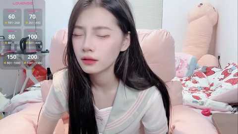 Video of an Asian girl with long black hair, wearing a white sailor-style top, lying on a pink bed with a plush toy and a red apple. Background includes a window and a digital display showing health metrics.