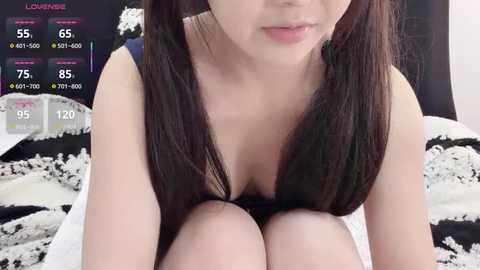 Video of an East Asian woman with long black pigtails, fair skin, wearing a black lace bra, sitting on a white and black bedspread in a dimly lit room.