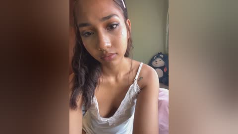 Video of a young woman with medium-brown skin and long black hair, wearing a white lace camisole, sitting indoors. Background features a stuffed toy panda and a bed with pink sheets.