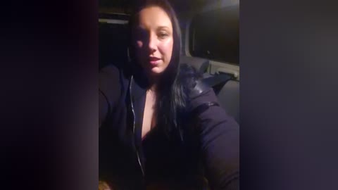 Media: A video of a young woman with long, dark hair, wearing a black jacket, seated in a dimly lit car interior. The image is slightly out of focus, with shadows dominating the background.