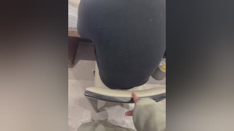 Video of a person in a tight-fitting black dress sitting on a white chair, with a hand holding a yellow towel, in a tiled bathroom.