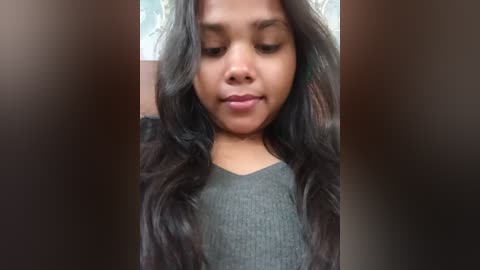 A video of a young girl with long, wavy black hair, wearing a grey ribbed sweater, gazing downward with a serene expression. The background is blurred.