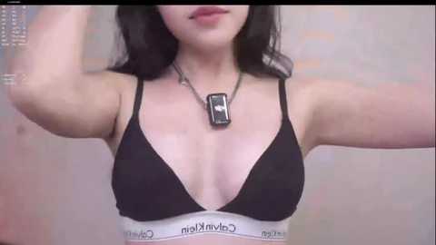 Video of a woman with fair skin, long dark hair, wearing a black Calvin Klein bralette, silver necklace, and a gray dog tag pendant.