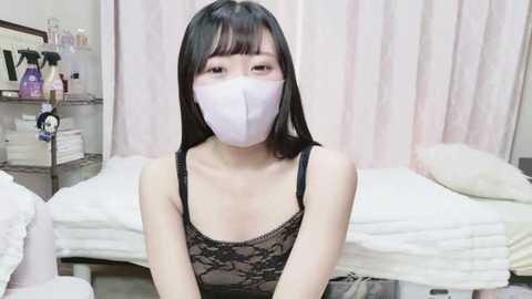 Media: A video of an Asian woman with long black hair, wearing a white face mask, black lace top, and seated on a white bed in a softly lit room.