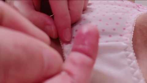 Video of a close-up of a child's hand with a small, sharp object, possibly a paperclip, delicately touching a white, polka-dotted garment, likely a piece of clothing, suggesting a curious, exploratory moment.