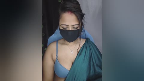 Video of a woman with medium skin tone, wearing a blue bra, black face mask, and teal blanket, standing in a dimly lit room.