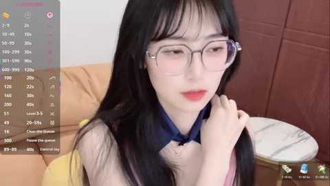 Video of an Asian woman with long black hair, wearing glasses and a yellow blouse, sitting on a couch in a dimly lit room.