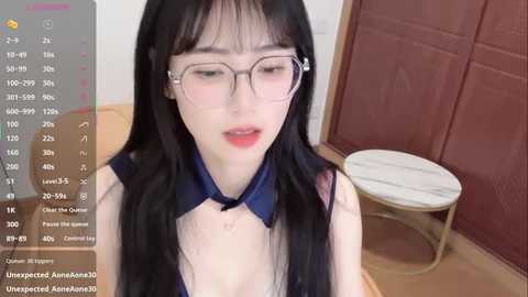 A video of a fair-skinned, slender Asian woman with long black hair and glasses, wearing a dark blue blouse and a bow tie, seated in a room with wooden floors and a brown door.