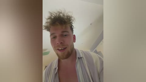 A video of a young, light-skinned man with curly brown hair and a beard, wearing a light blue shirt, smiling. The background is a plain, beige room.