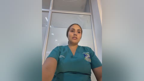 Media: Video of a Latina nurse with medium skin tone, brown hair tied back, wearing teal scrubs with \"Jewish Hospital\" logo, standing in a white room with frosted glass windows and ceiling lights.