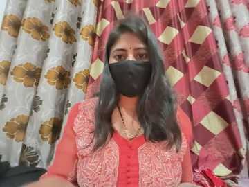 Video of a South Asian woman with medium brown skin, long black hair, and a black face mask, wearing a red and gold embroidered blouse, standing in front of patterned curtains with floral and diamond motifs.