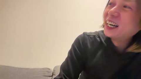 Video of a smiling woman with short, light brown hair, wearing a dark gray long-sleeve shirt, sitting on a beige sofa against a plain beige wall.