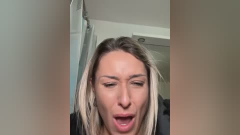 A video of a blonde woman with fair skin, wearing a black top, in a bathroom, looking shocked and opening her mouth wide.
