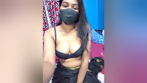 A video of a South Asian woman with medium skin tone, long black hair, wearing a black bra and face mask, seated in a messy bedroom with blue and red walls.