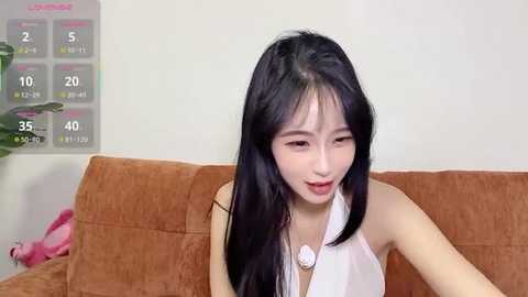 Video of an Asian woman with long black hair, fair skin, and a slim figure, wearing a white halter top, sitting on a brown couch, looking down, with a pink stuffed toy and a calendar in the background.