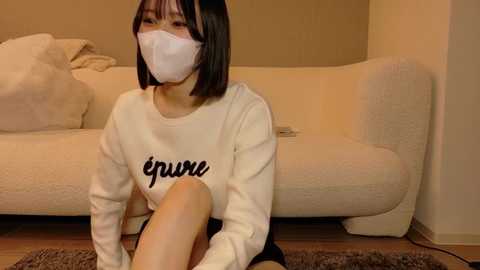 Video of an Asian woman with short black hair, wearing a white face mask and a white sweater with \"Euphoria\" printed in black, sitting on a brown rug in front of a beige couch.