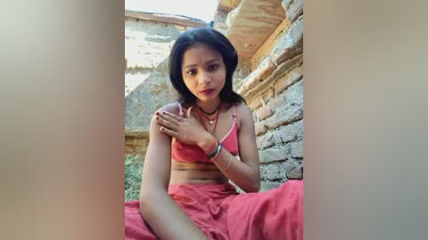 Video of a young woman with medium skin tone and black hair, wearing a red sari, sitting on a red cloth in an old, weathered stone structure.