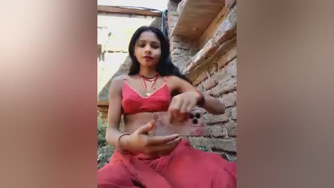 Video of a young woman with medium brown skin, long black hair, wearing a red bra and pants, sitting against a brick wall, holding a glass of water.