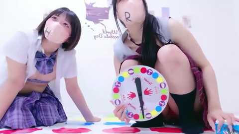 Video of two young Asian women in schoolgirl outfits, playing Twister on a colorful mat, with \"R\" written on their faces.