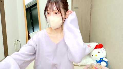 Video of a young East Asian woman with pale skin, wearing a light purple sweater and a white face mask, adjusting her hair in a bedroom. Background features a white door and a Hello Kitty plush toy on a bed.