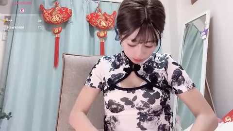 Video of an Asian woman with straight black hair, wearing a traditional Chinese cheongsam with black floral pattern, standing in front of a mirror with red and gold decorations.