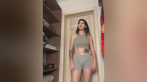 Video of a slender woman with medium-length, wavy brown hair, wearing a light green sports bra and high-waisted shorts, standing in a modern, well-organized bedroom with wooden shelves and a red floral curtain.