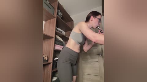 A video of a Caucasian woman with short brown hair, glasses, wearing a gray sports bra and leggings, using a pink lipstick in a bathroom with wooden shelves and a beige door.