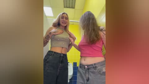 Video of two women in a bathroom, one wearing a gray crop top and high-waisted jeans, the other in a pink tank top and baggy pants, both smiling and touching each other's stomachs.