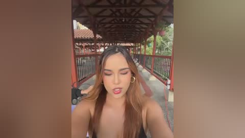 Video of a young woman with long, wavy hair and light skin, wearing a black top, taking a selfie under a red metal bridge.