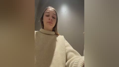 A video of a young Caucasian woman with straight, light brown hair, wearing a beige, ribbed-knit sweater, looking down at a book in a dimly lit, plain room with neutral-colored walls.