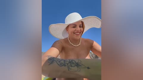 Video of a smiling, topless woman with light skin and tattoos, wearing a large white sunhat, standing in front of a clear blue sky.