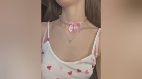Video of a young woman with fair skin, long brown hair, wearing a white spaghetti-strap tank top with red heart patterns, pink bow choker, and silver necklace.
