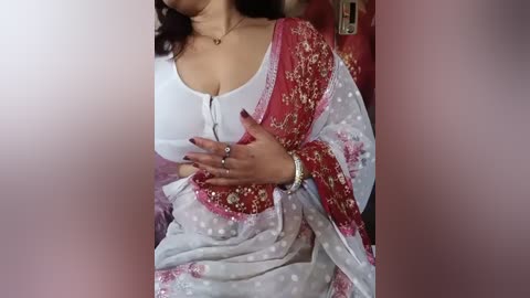 Video of a woman with medium skin tone, wearing a white blouse and a red and white floral sari, hands pressed against her chest, with a blurred background.
