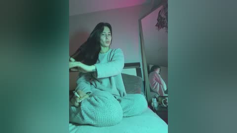 A video shows a young woman with long dark hair, sitting on a bed, wearing a light-colored sweater and pants, brushing her hair in a dimly lit room with a mirror.
