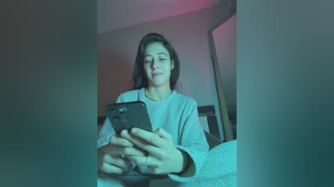 A video of a young woman with long dark hair, wearing a light gray sweater, sitting in a dimly lit room, focused on a smartphone.