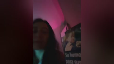 A blurry, dimly lit video captures a person with dark hair and a blue shirt, partially obscured by another figure. The background features a pinkish hue and a poster with Spanish text.