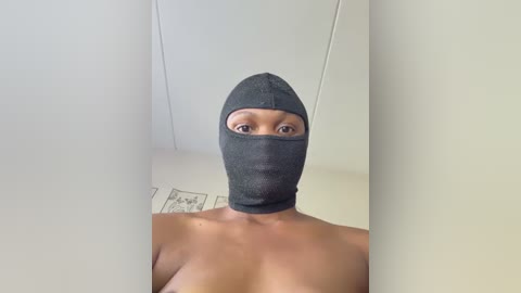Video of a topless woman with light brown skin and medium-sized breasts wearing a grey balaclava, standing in a plain, white-walled room with a tiled floor.