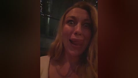Video of a blonde woman with shoulder-length hair, wearing a white top, making a playful face, indoors with dim lighting and string lights in the background.