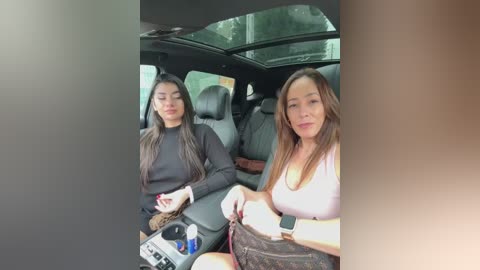 Video of two young women in a car. One wears a black sweater, the other a white top, both with long hair. They sit in the backseat, looking ahead.