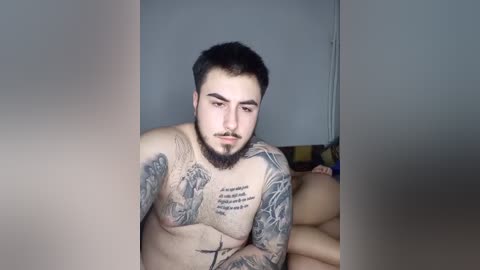 Video of a shirtless man with a full beard, dark hair, and tattoos on his chest and arms, sitting on a bed in a dimly lit room.