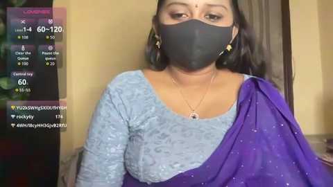 A video of an Indian woman in a grey lace blouse and purple saree, wearing a black face mask, with a background of beige walls and a partially visible green plant.
