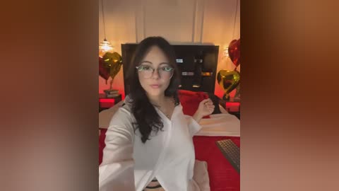 A video of a woman with long, dark hair and glasses, wearing a white blouse, sitting at a table with red and white tablecloths, surrounded by heart-shaped balloons. The background features a TV and a warm, dimly-lit room.