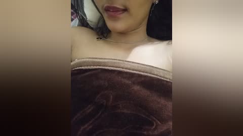 Video of a woman's upper body, partially covered by a brown towel, lying on a bed with a pillow. She has light skin and wears a small necklace.