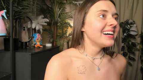 Video of a smiling young woman with fair skin, brown hair, and tattoos on her collarbone, wearing a necklace, in a room with plants, a shelf with toys, and a mirror.