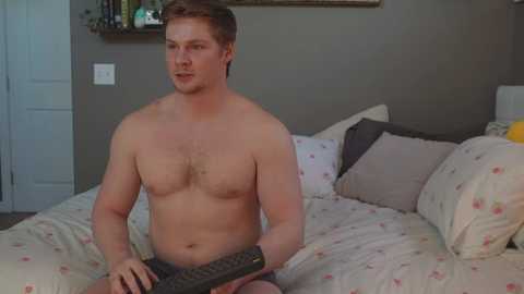 Media: Video of a shirtless, muscular white man with short brown hair, sitting on a bed with floral sheets, holding a TV remote.